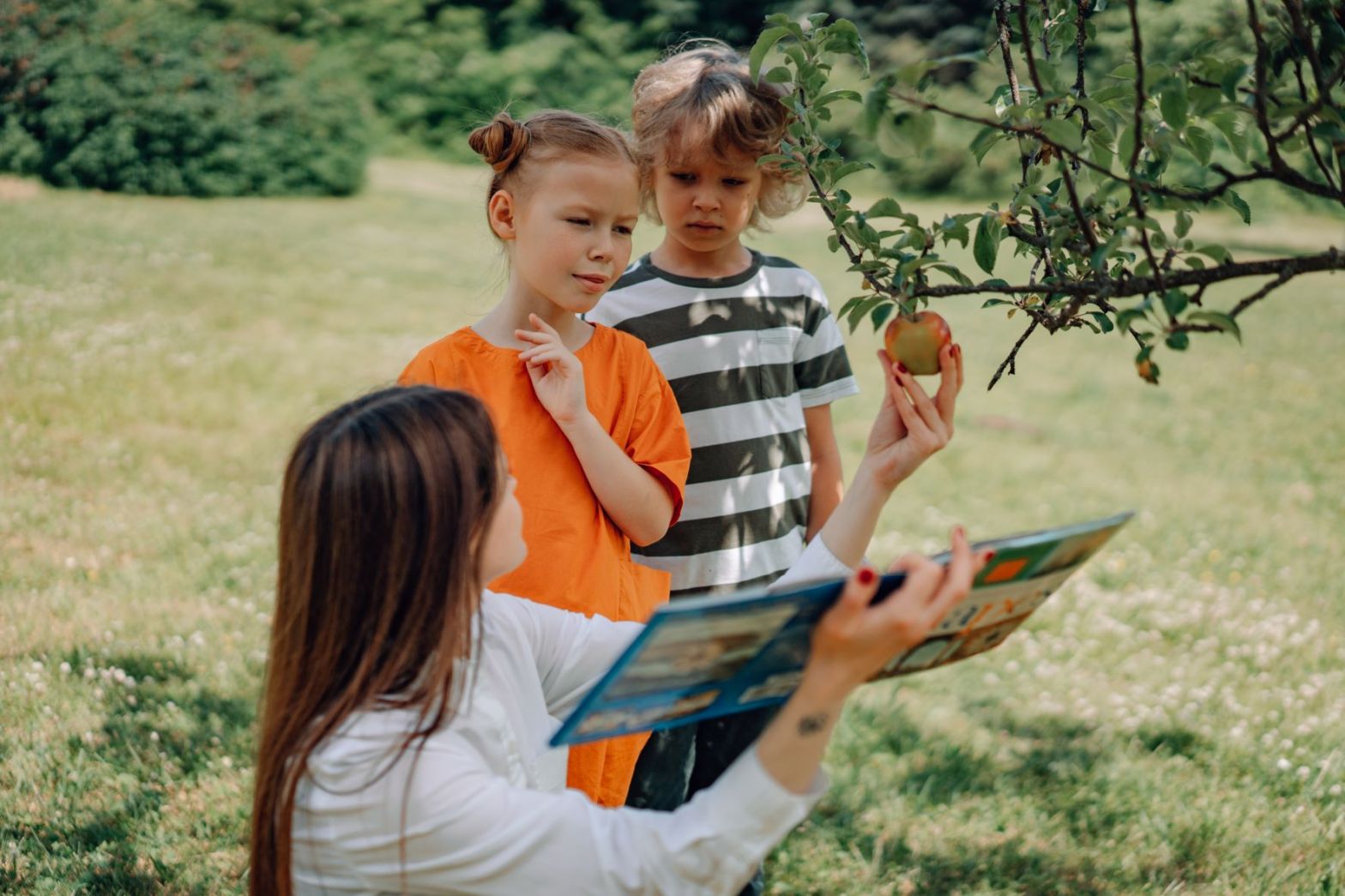 two kids reading outdoors with a woman holding an apple